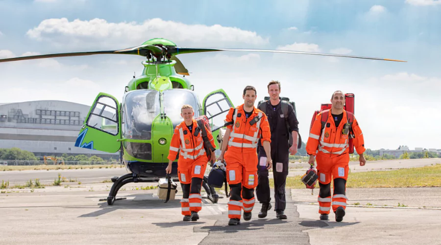 Four air ambulance paramedics waking in front of a green helicopter wearing orange high-visibility jumpsuits.