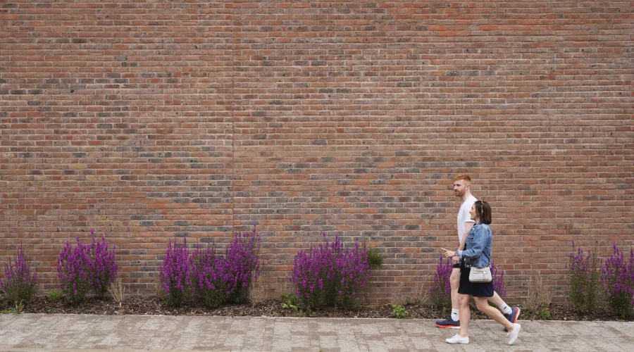 Couple walking in causal summer clothing along street in Brabazon showcasing purple spring flowers in front of brick wall.