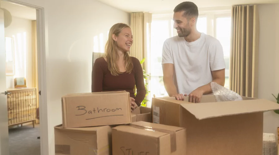 Couple inside new Dials Apartment living room laughing while unpacking their belongings out of brown boxes.