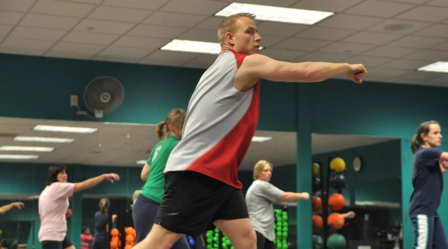 Small group aerobics fitness class taking place in a green sports room with focus on male in sports singlet.